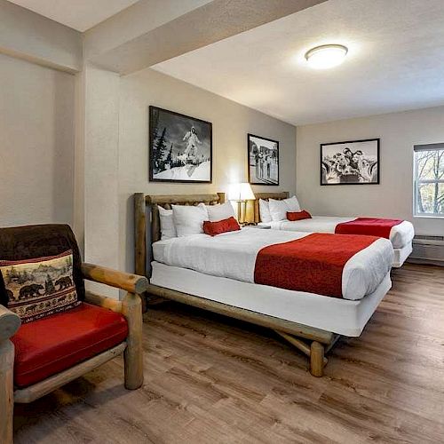 A cozy hotel room features two beds with red throws, a wooden armchair, framed art on the walls, a TV on a dresser, and a large window.