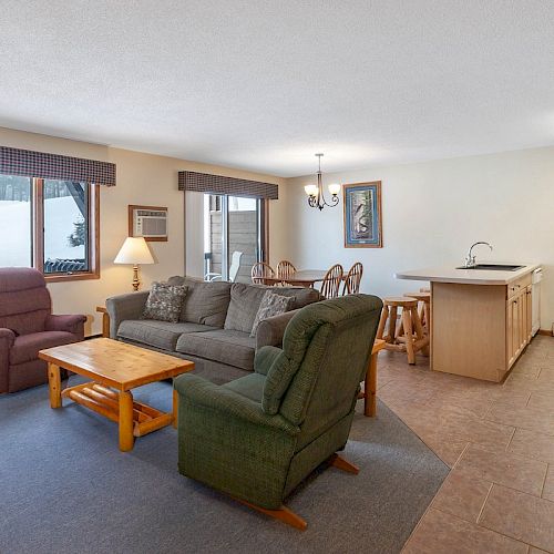 The image shows a cozy living area with a sofa, two armchairs, a coffee table, a dining table, and a kitchen with an island and stools.