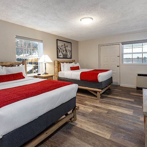 A hotel room with two double beds featuring red and white bedding, a TV on a wooden stand, a lamp by the window, and art on the wall, is shown.