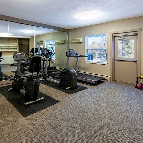 This image shows a small gym with exercise equipment including a treadmill, cycling machines, and weights, along with a large mirror.