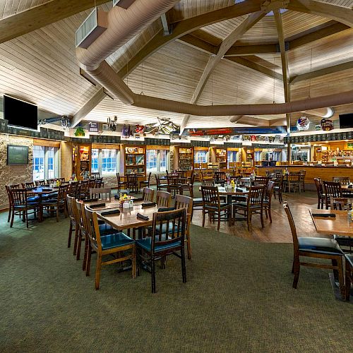 The image shows the interior of a spacious, rustic-style restaurant with wooden furnishings, several tables and chairs, a bar area, and a high beamed ceiling.