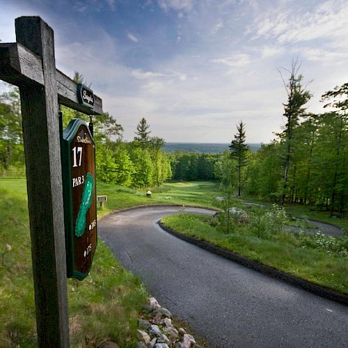 The image shows a sign for hole 17 at a golf course, with a winding path leading downhill and lush greenery on both sides, under a partly cloudy sky.