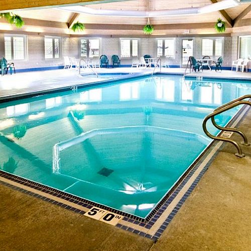 The image shows an indoor swimming pool with clear water, ladders at either end, lounge chairs around the perimeter, and potted plants hanging.