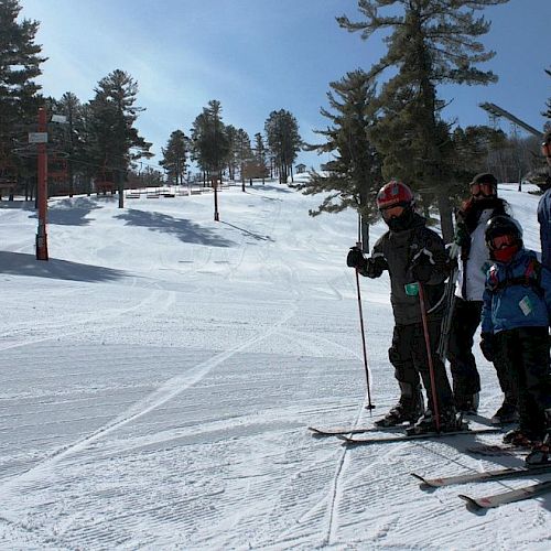 A group of people, equipped with skis and poles, stand on a snowy slope beneath a clear sky, surrounded by trees, near a ski lift.