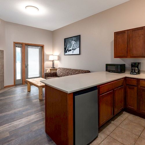 The image shows a cozy living area with a kitchen, featuring wooden cabinets, a small fridge, a sofa, a wall-mounted TV, and a fireplace.