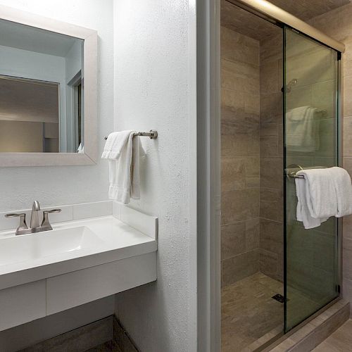 A bathroom with a sink, mirror, towel rod, glass-enclosed shower, and a toilet with folded towels above it is shown.