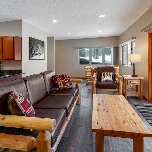 A cozy living room features a dark brown sofa, wooden coffee table, and sliding glass doors opening to a snowy patio, with a kitchen in the background.