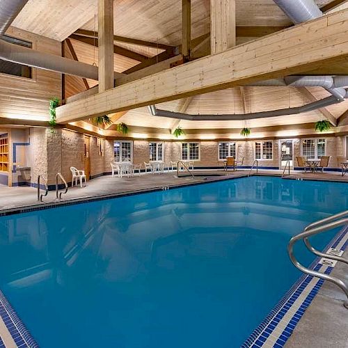 An indoor swimming pool with blue water, surrounded by wooden beams and lounge chairs, located in a well-lit, modern facility.