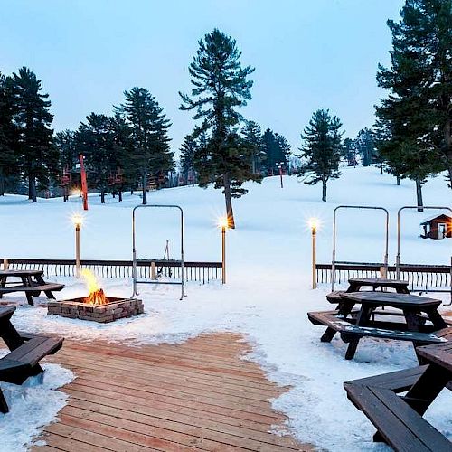 An outdoor snowy setting with picnic tables, a fire pit, tiki torches, and pine trees in the background, creating a cozy atmosphere.