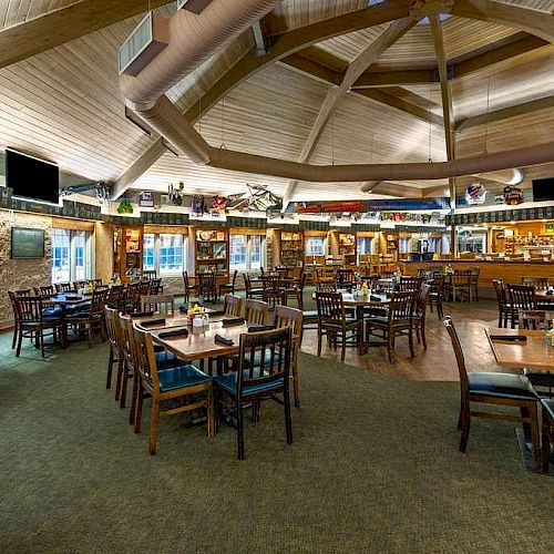 A spacious restaurant interior with wooden tables and chairs, a central bar area, and a rustic ceiling design. The space is well-lit and inviting.