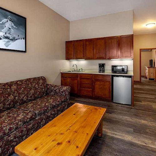 The image shows a furnished living area with a floral couch, wooden coffee table, and kitchenette, featuring a mini-fridge and microwave.