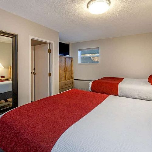 The image shows a hotel room with two double beds, red accents, a large mirror, a nightstand, a lamp, and a wooden dresser.