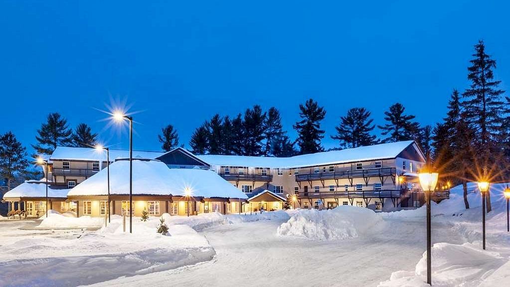A cozy lodge surrounded by snow, warmly lit by street lamps, with tall pine trees in the background under a clear twilight sky.