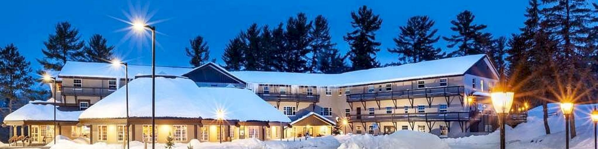 A cozy lodge surrounded by snow, warmly lit by street lamps, with tall pine trees in the background under a clear twilight sky.