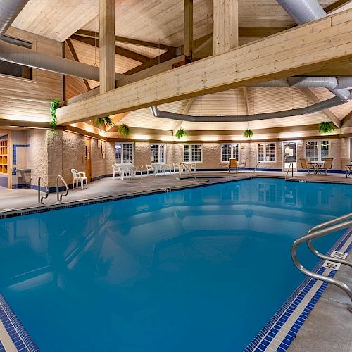 This image shows an indoor swimming pool in a well-lit space with wooden ceiling beams, pool chairs, and plants around the pool area.