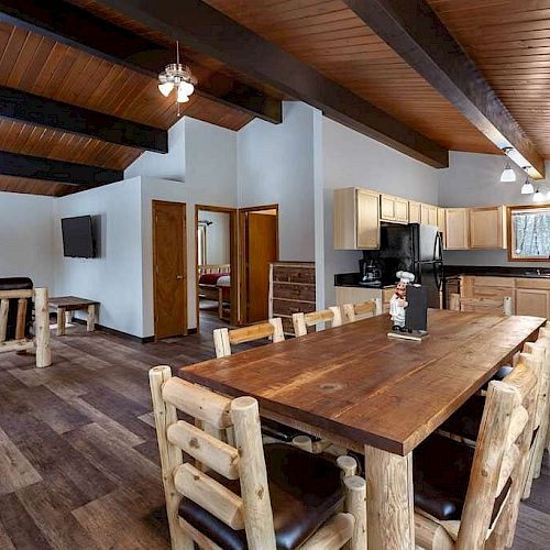 The image shows a rustic-style open-concept living space with a wooden dining table and chairs, a cozy lounge area, and a kitchen.