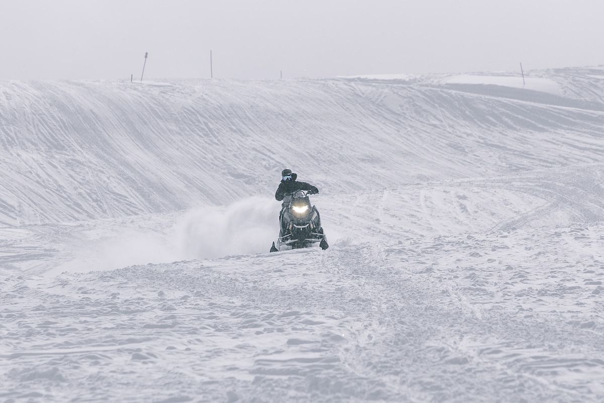 A person is riding a snowmobile in a snowy landscape, creating a trail in the snow. The surroundings are covered in snow with low visibility.