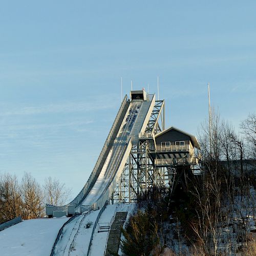 The image shows a snow-covered ski jump ramp with a structure at the top, surrounded by trees, under a clear blue sky.