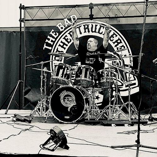 A three-member band performs on stage with guitars and drums, in front of a sign that reads 