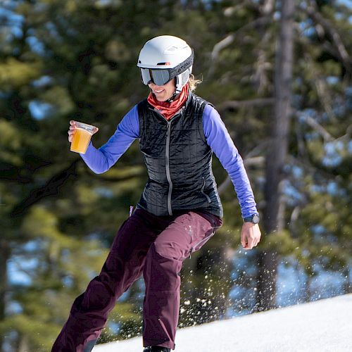 A skier in protective gear is descending a snowy slope while holding a beverage cup and smiling.