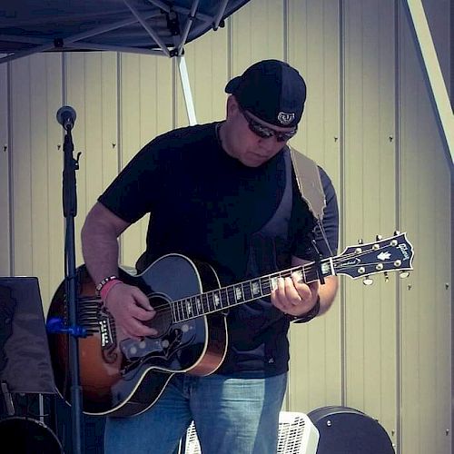 A person wearing sunglasses and a backward cap plays the guitar in front of a microphone stand, with a speaker and equipment in the background.