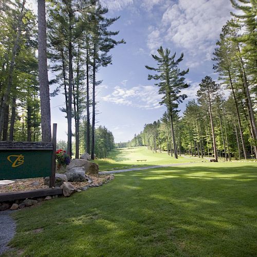 The image shows a scenic golf course surrounded by tall trees and a sign with a logo on the left side, leading to a fairway under a blue sky.