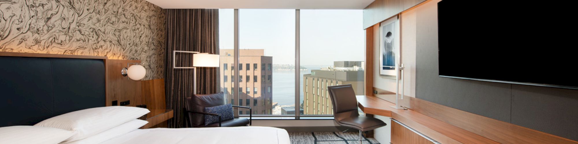 Modern hotel room with a large bed, wall-mounted TV, desk, chair, lounge chair, and window with a city view in daylight.