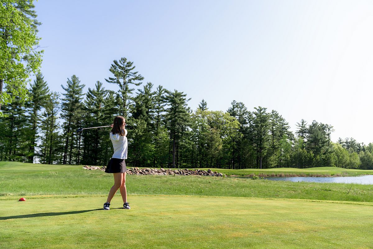 A person is swinging a golf club on a lush green course with trees and a small pond in the background.