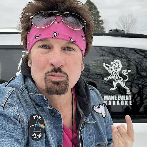 A person with a pink headband and sunglasses making a gesture in front of a vehicle with "Mane Event Karaoke" and a lion decal on the window.