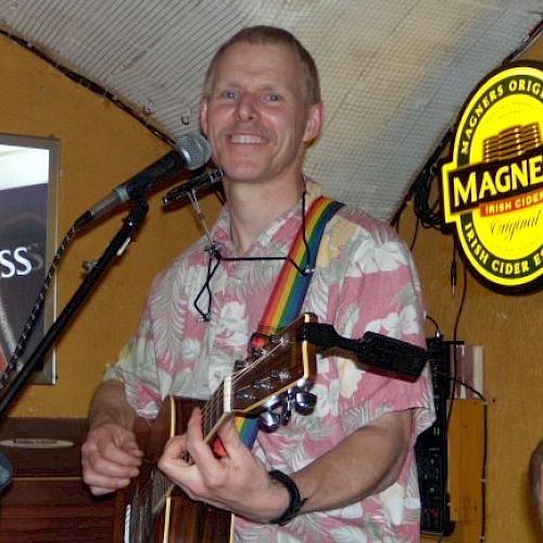 A person smiling and playing guitar on stage, wearing a floral shirt with a rainbow strap, a microphone in front. Magner's cider sign in the background.