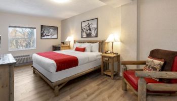 This image shows a cozy bedroom with a double bed, red accents, wooden furniture, two lamps, framed photos, and a window with a scenic view.