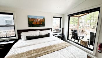 A cozy bedroom with a large bed, windows, and a sign saying "Adventure Awaits" above the bed, overlooking an outdoor deck area.