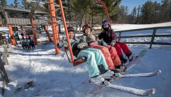 Three people in ski gear sitting on a ski lift, smiling, with a snowy landscape and trees in the background, ready for skiing adventures.