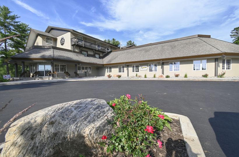 A building with multiple stories and a large parking lot in front, featuring a rock and flower bed in the foreground, under a clear sky.