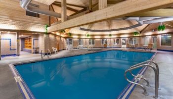 This image shows an indoor pool with clear blue water, surrounded by lounge chairs, plants, and wooden beams under a high ceiling.