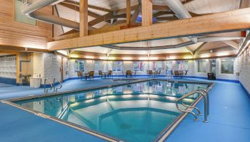 An indoor swimming pool with blue flooring, surrounded by tables, chairs, and wooden beams in a spacious room.