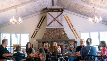 People dining and socializing in a cozy, rustic restaurant with a stone fireplace and ski decor, under a vaulted ceiling with chandeliers.