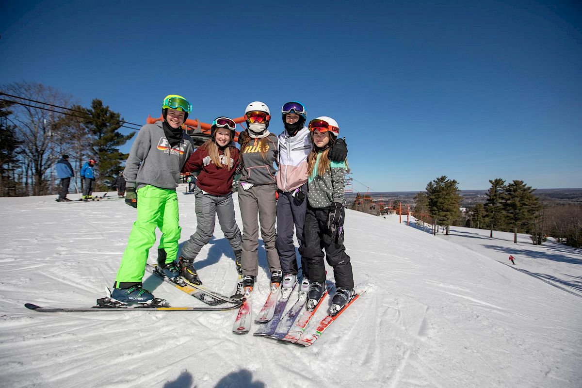 A group of five people in ski gear posing on a snowy slope with trees and a clear sky in the background.