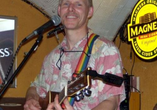 A person is playing an acoustic guitar and singing at a venue. He is smiling, wearing a floral shirt with a rainbow guitar strap.