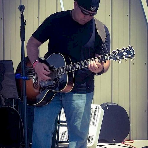 A person is playing an acoustic guitar, standing next to a microphone stand, wearing a black shirt, blue jeans, and a cap, outside by a building.