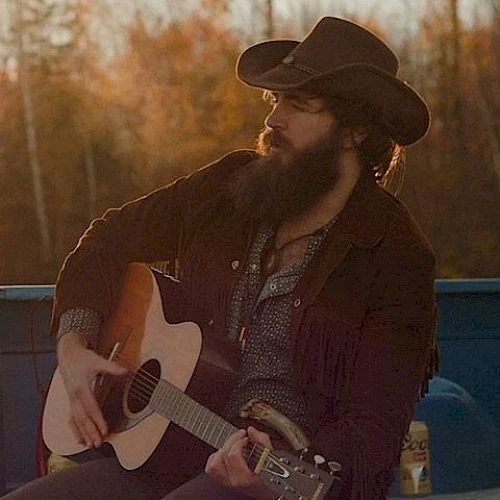 A person with a beard, wearing a cowboy hat, is playing an acoustic guitar outdoors. There is a beverage can to the right.