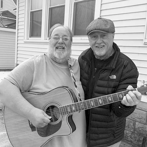 Two elderly men are smiling, standing outside a house. One is holding a guitar while the other has an arm around him, also holding the guitar.