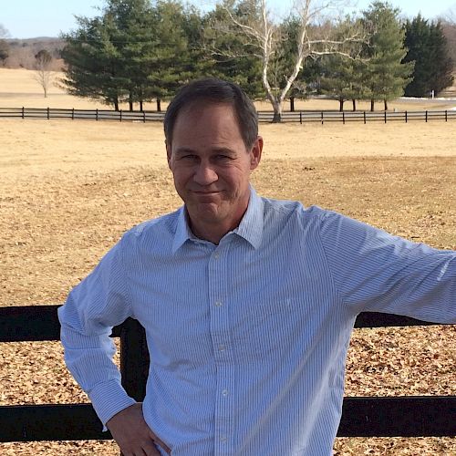 A person is standing outdoors next to a fence, wearing a light blue shirt. The background features an open field with trees, under a bright sky.