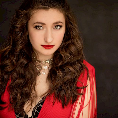 A woman with wavy hair and red lipstick is wearing a red outfit and a decorative necklace, standing against a dark background.