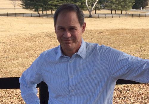 A person in a light-colored shirt stands against a wooden fence in a rural area with trees and an open field in the background.