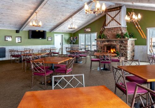 The image shows a cozy dining area with wooden tables, chairs, a stone fireplace, and rustic decor, including chandeliers and a TV.