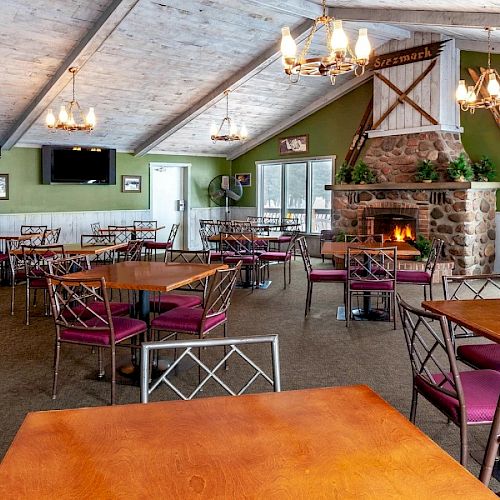 The image shows a cozy dining area with wooden tables, chairs, a stone fireplace, and rustic decor, including chandeliers and a TV.