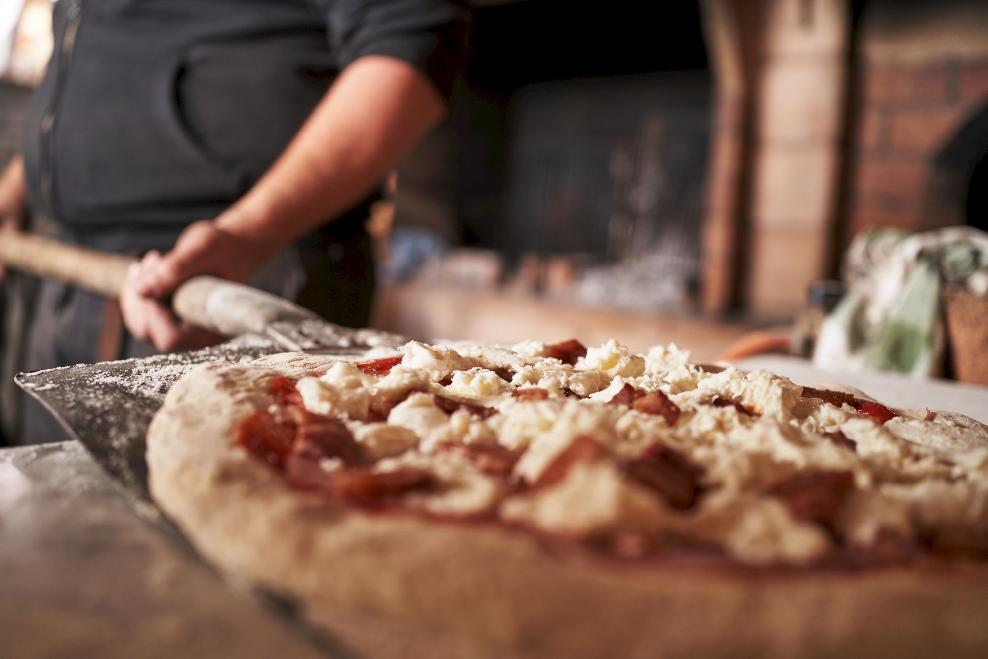 A person is preparing to place a pizza, topped with cheese and tomato sauce, into a brick oven using a pizza peel.