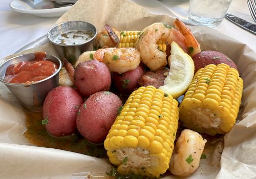 The image shows a seafood boil platter with shrimp, corn on the cob, red potatoes, a lemon wedge, and side sauces on a paper-lined tray.