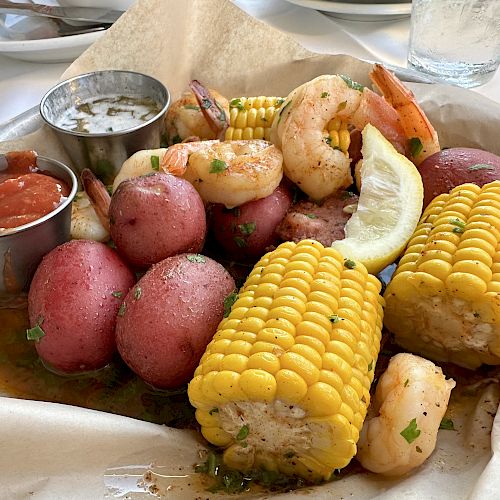 The image shows a seafood boil platter with shrimp, corn on the cob, red potatoes, a lemon wedge, and side sauces on a paper-lined tray.